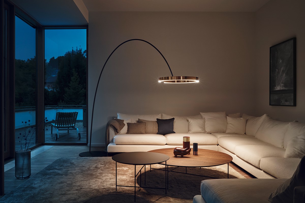 Abendstimmung im Wohnzimmer mit grossem, weissem Ecksofa und vielen Kissen. Über dem Sofa steht eine bogenförmige Stehleuchte mit ringförmigem Lichtkörper. Davor zwei niedrige Couchtische auf einem weichen Teppich. Links eine bodentiefe Glasfront mit Blick auf Terrasse und Liegestuhl in der Dämmerung.