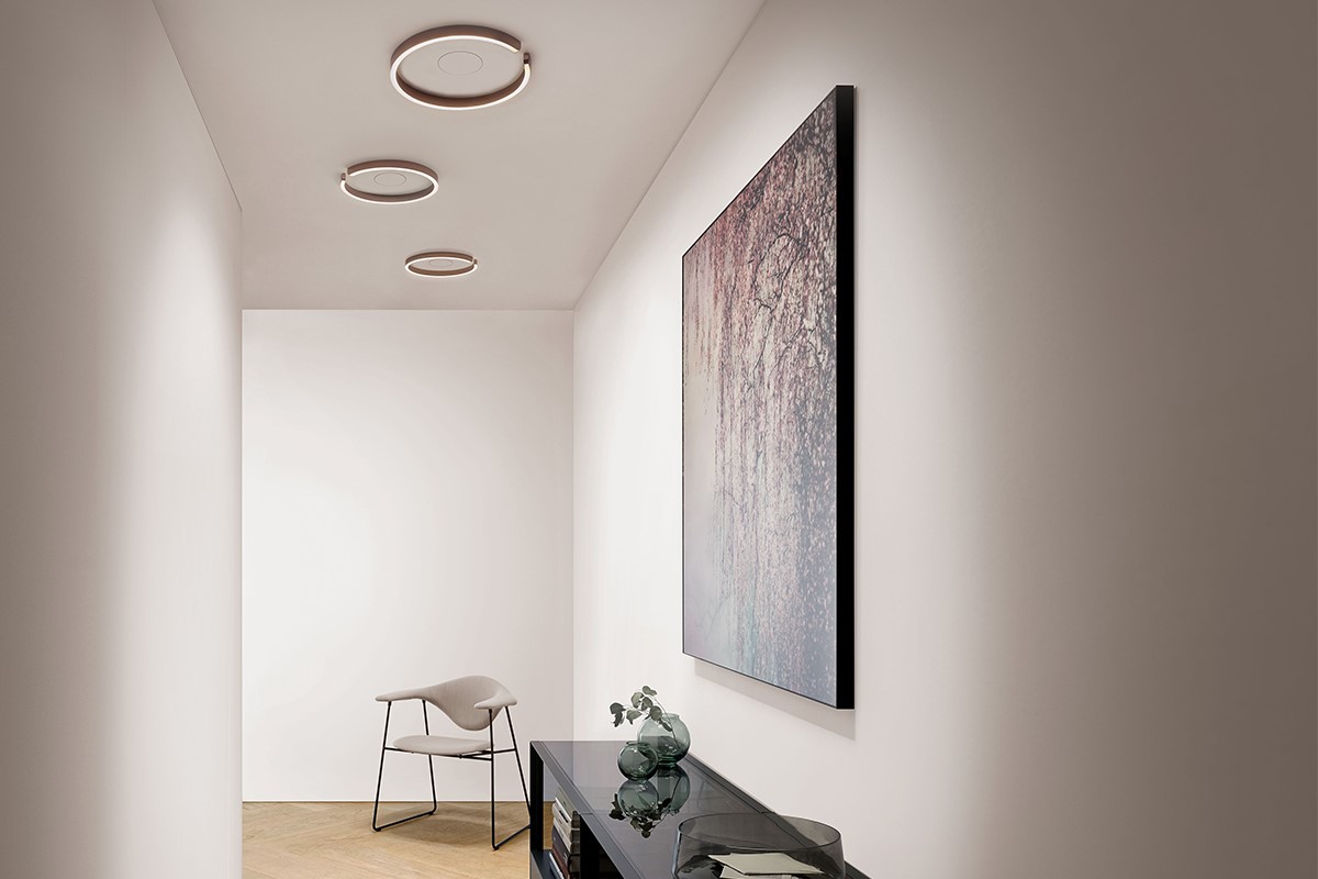 Heller Flur mit weissen Wänden und drei runden Deckenleuchten. Rechts hängt ein grossformatiges Kunstbild über einem schwarzen Sideboard mit Glasvasen. Am Ende des Flurs steht ein leichter Stuhl mit Metallgestell auf hellem Holzboden.