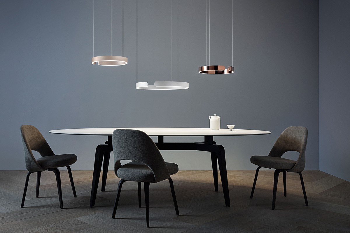 Minimalistischer Essbereich mit ovalem, weissem Esstisch und schwarzem Gestell. Drei gepolsterte Stühle in Dunkelgrau stehen um den Tisch. Darüber hängen drei Pendelleuchten in Ring und Line Form, teils in Kupferoptik. Ruhige Kulisse mit graublauer Wand und dunklem Holzboden.