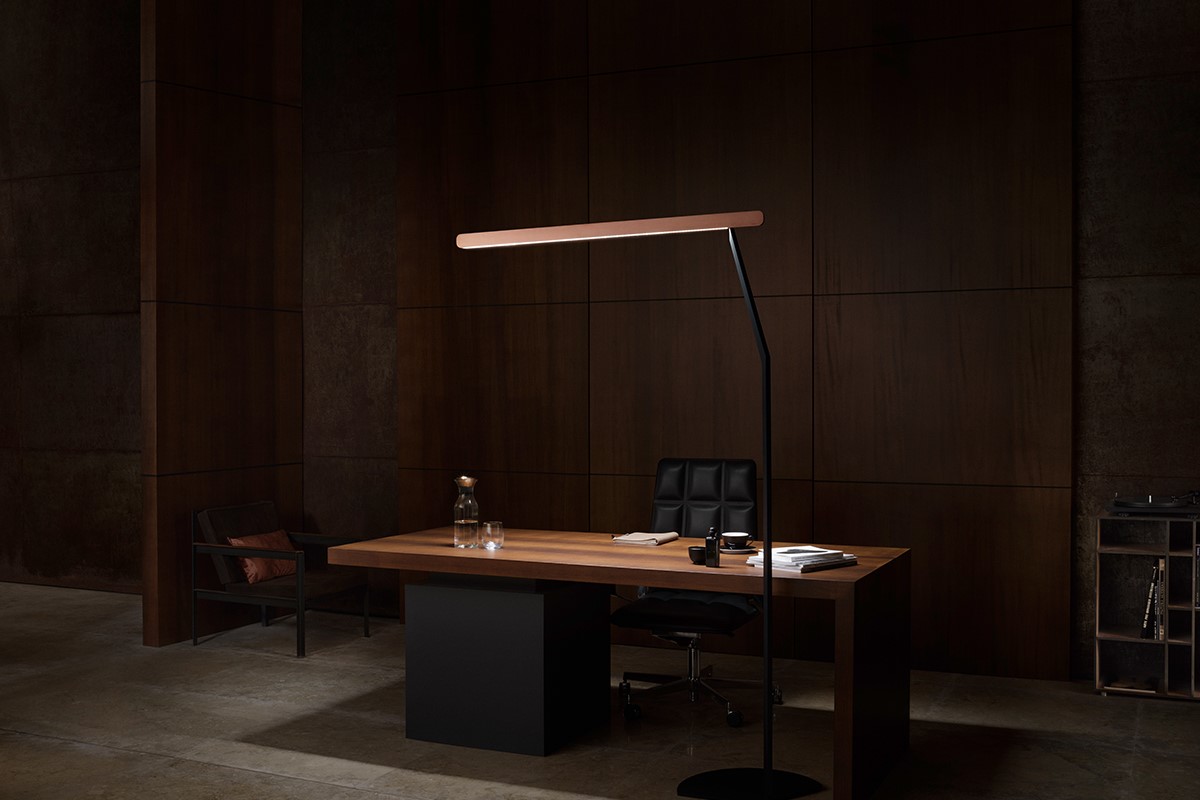 Dunkles Office Setting mit grossem Holztisch vor wandfüllender Holzvertäfelung. Hinter dem Tisch steht ein schwarzer Drehstuhl, links ein niedriger Sessel. Eine schlanke Stehleuchte mit horizontalem Lichtbalken setzt einen warmen Akzent. Auf dem Tisch liegen Unterlagen, dazu Karaffe, Gläser und kleine Accessoires. Rechts steht ein schmales Regal.