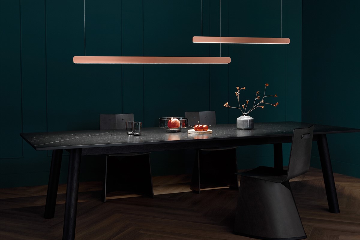 Dunkler Essbereich mit langem Tisch in schwarzer Steinoptik und schwarzen Stühlen. Vor einer tiefgrünen Wand hängen zwei schlanke Pendelleuchten in warmem Rosa. Auf dem Tisch stehen Gläser, eine Schale mit Äpfeln und eine Vase mit Zweigen. Dunkler Holzboden rundet das reduzierte Setting ab.
