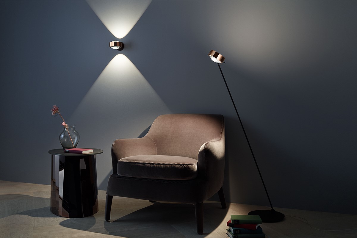 Leseecke mit gepolstertem Sessel in Taupe vor dunkelblauer Wand. Eine Wandleuchte erzeugt einen X förmigen Lichtkegel, daneben steht eine schlanke Stehleuchte mit Metallkopf. Links ein kleiner Beistelltisch mit Glasvase und Buch. Am Boden liegen weitere Bücher.