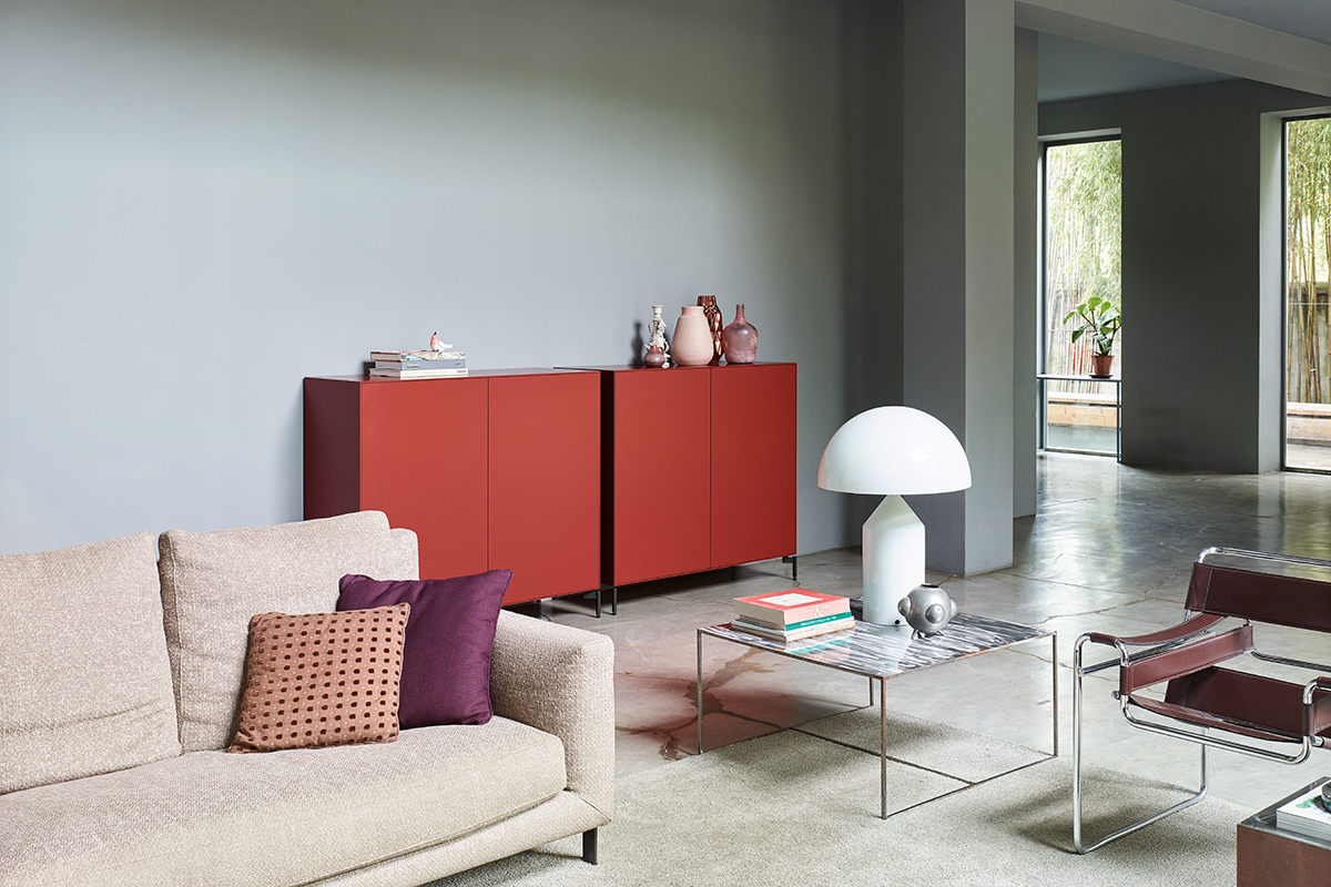 Moderner Wohnbereich mit hellem Sofa und Kissen in Beige und Violett. An der Wand stehen zwei rote Sideboards mit Vasen und Büchern. Davor ein niedriger Tisch mit Marmorplatte und weisser Tischleuchte. Rechts ein Sessel mit Chromgestell. Betonboden und graue Wände sorgen für eine klare, reduzierte Atmosphäre.