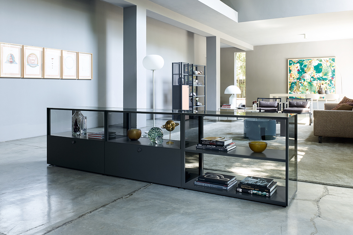 Grosszügiger Showroom mit minimalistischer Einrichtung. Im Vordergrund steht ein langes Sideboard mit schwarzem Rahmen und Glasflächen, gefüllt mit Büchern und Dekoobjekten. Im Hintergrund Lounge Möbel, Steh und Tischleuchten sowie ein farbiges Kunstwerk an der Wand. Helle Wände, Betonboden und viel Tageslicht prägen den Raum.