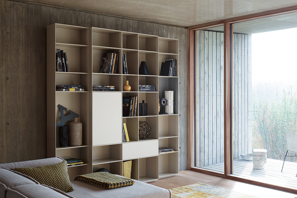 Wohnraum mit grossem, hellem Regal mit offenen Fächern, Büchern und Dekoobjekten. Daneben bodentiefe Schiebetüren mit Holzrahmen und Blick auf eine gedeckte Terrasse. Sichtbetonwände und heller Holzboden schaffen eine reduzierte, moderne Atmosphäre.