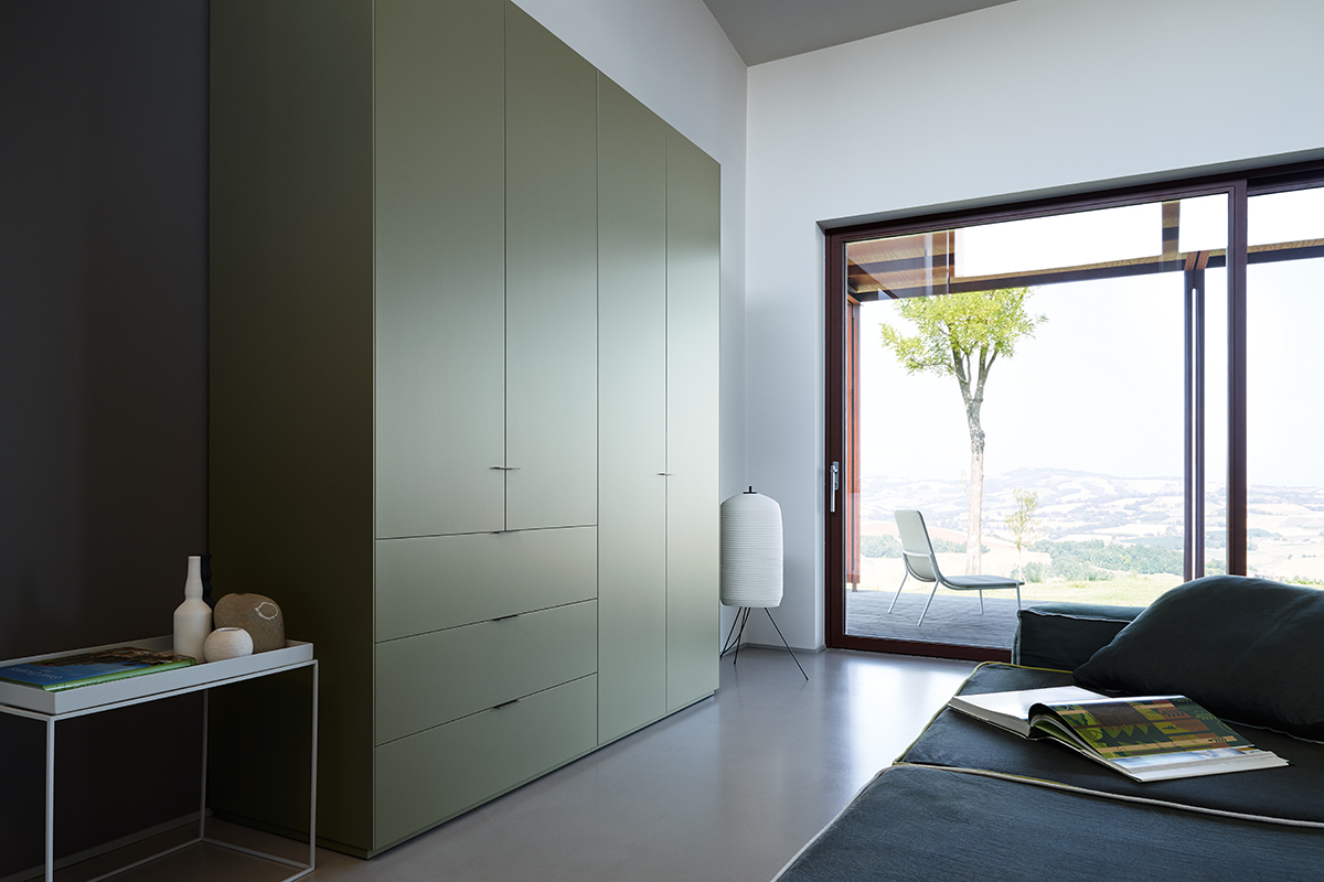 Minimalistischer Wohnraum mit deckenhohem Einbauschrank in Salbeigrün und flächenbündigen Fronten. Links eine dunkle Akzentwand mit kleinem Beistelltisch und Keramikobjekten. Rechts eine grosse Schiebetür zur Terrasse mit Stuhl, Baum und Weitblick in die Landschaft. Im Vordergrund ein dunkles Sofa mit aufgelegtem Magazin. Eine Stehleuchte steht neben dem Fenster.