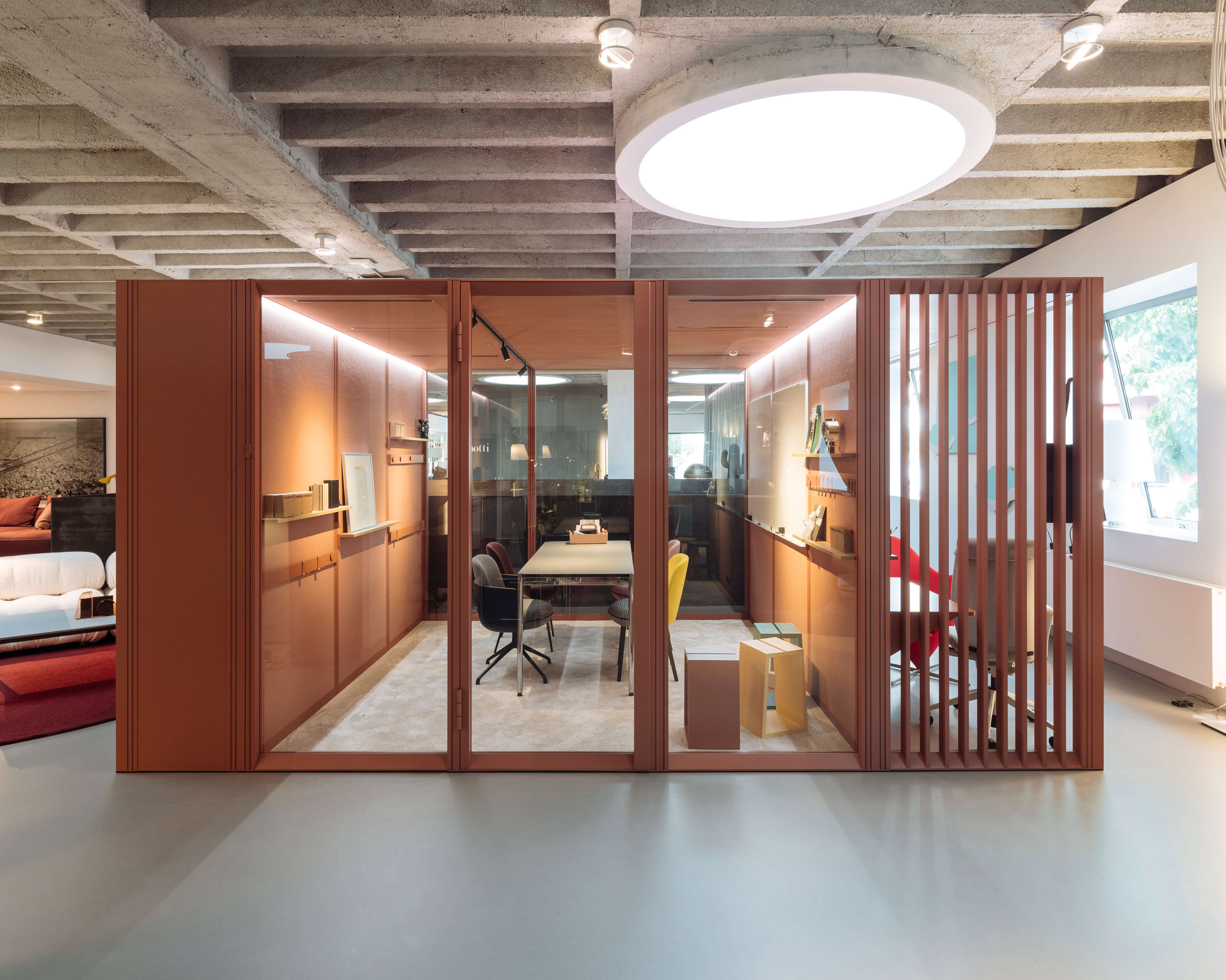 Büro Showroom mit modularer Raum-in-Raum Box in Terrakotta. Hinter Glasfronten sind ein Meetingtisch mit Stühlen, Whiteboards und Regale sichtbar. Rechts trennt ein Lamellen Element einen zweiten Bereich ab. Beton Decke, grosse runde Deckenleuchte und Fenster sorgen für offene Atmosphäre.