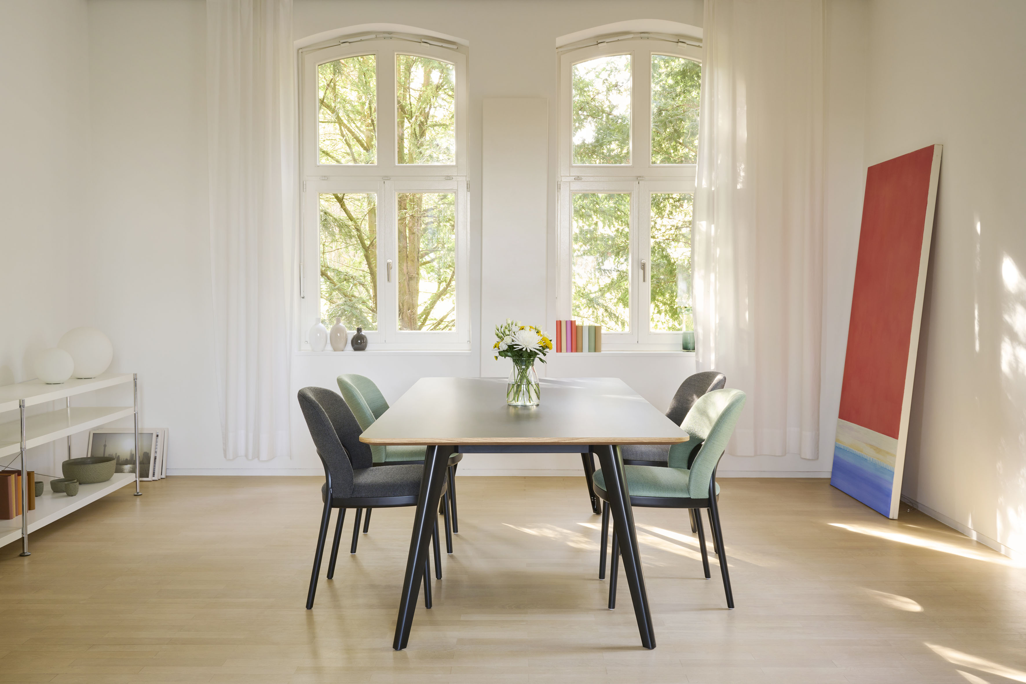 Heller Essbereich mit rechteckigem Holztisch und vier gepolsterten Stühlen in Grau und Mint. Grosse Rundbogenfenster mit weissen Vorhängen, Vase mit Blumen auf dem Tisch, farbiges Bild an der Wand, heller Holzboden.