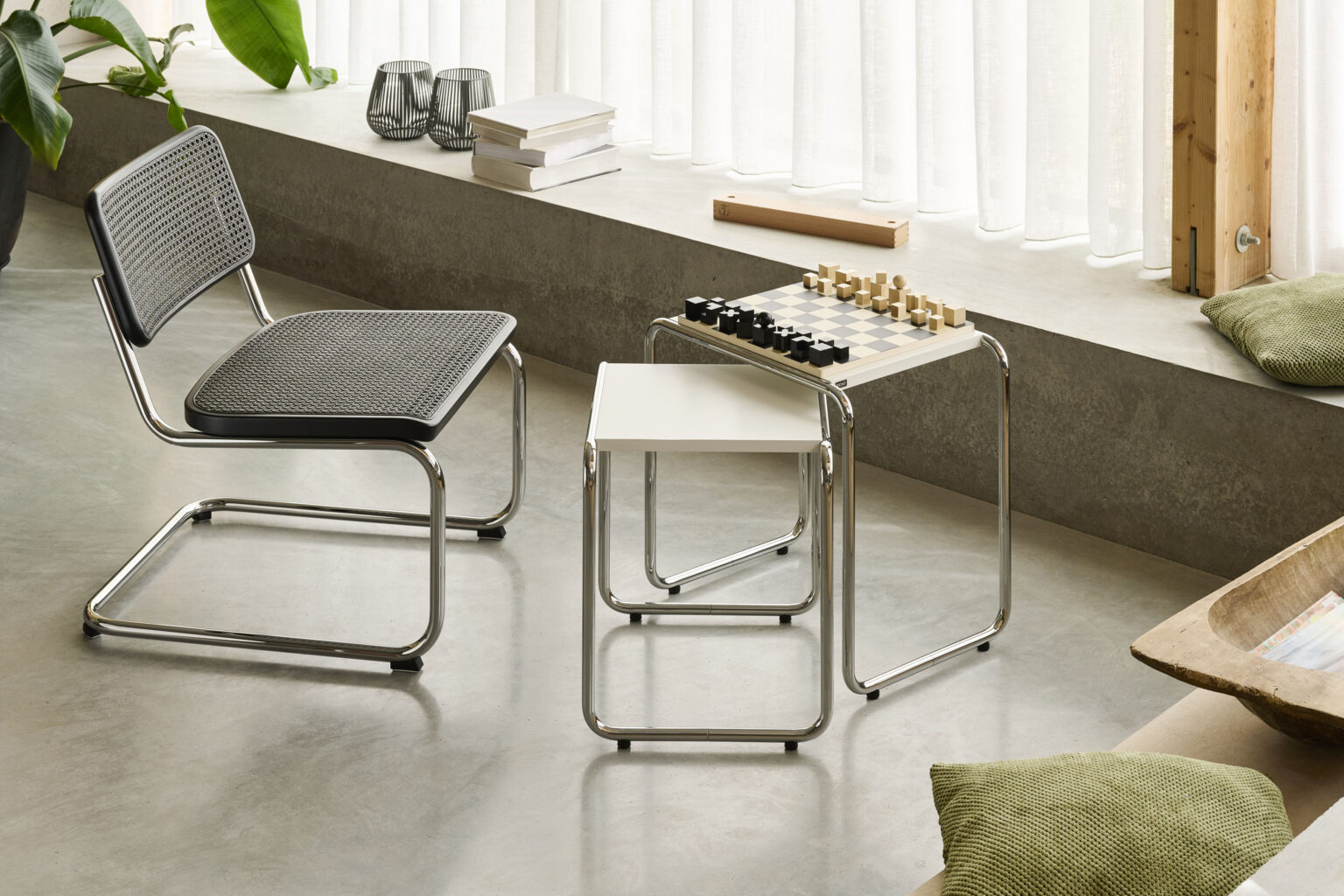 Helles Wohnambiente mit Freischwinger in Geflecht. Zwei stapelbare Beistelltische mit verchromtem Rohrgestell. Darauf ein Schachbrett mit Figuren. Betonboden und bodentiefe Fenster mit weissen Vorhängen. Kissen und Deko im Hintergrund. Minimalistische Atmosphäre.