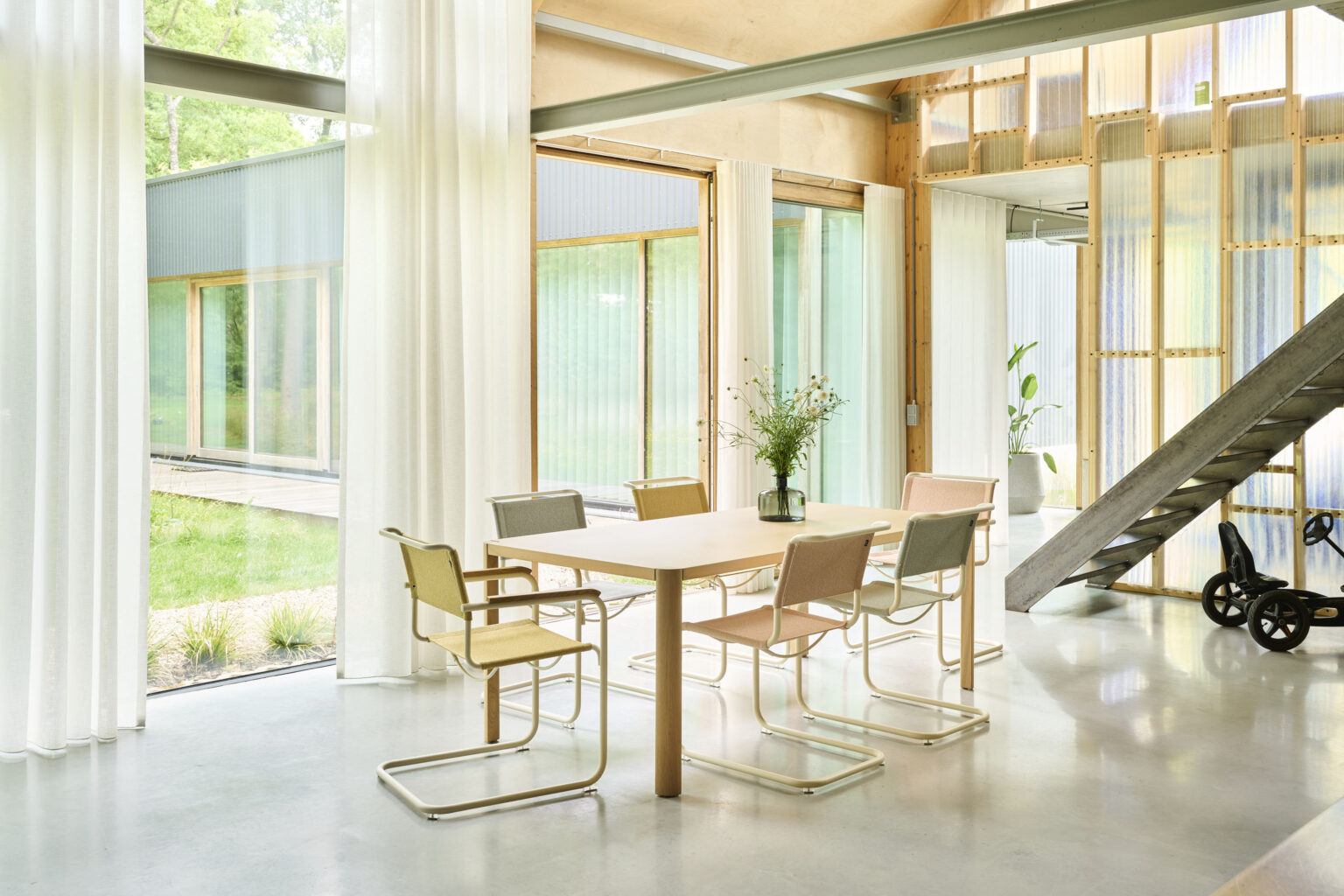 Heller Essbereich mit bodentiefen Vorhängen und Sichtbetonboden. Heller Holztisch mit sechs Freischwingern in Pastell und Chrom. Hintergrund mit transluzenten Wandpanelen, Pflanzen und Metalltreppe.