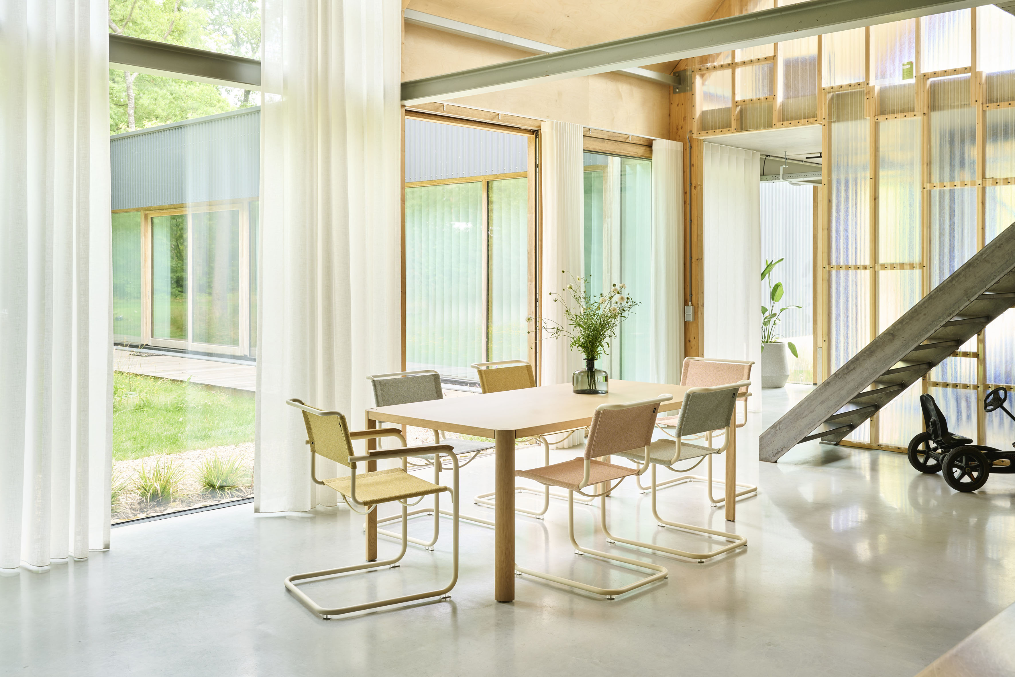 Heller Essbereich mit bodentiefen Vorhängen und Sichtbetonboden. Heller Holztisch mit sechs Freischwingern in Pastell und Chrom. Hintergrund mit transluzenten Wandpanelen, Pflanzen und Metalltreppe.