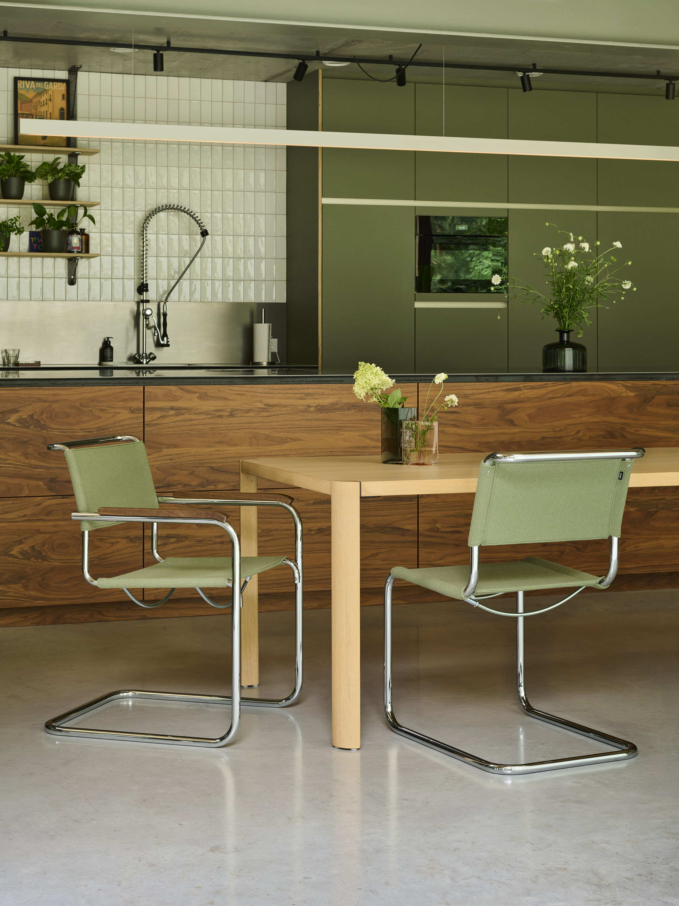 Küche mit grünem Schrankensemble und Holzfronten. Heller Holztisch mit zwei Freischwingern in Grün und Chrom. Offene Regale mit Pflanzen, weisse Fliesen und Profi Armatur im Hintergrund.