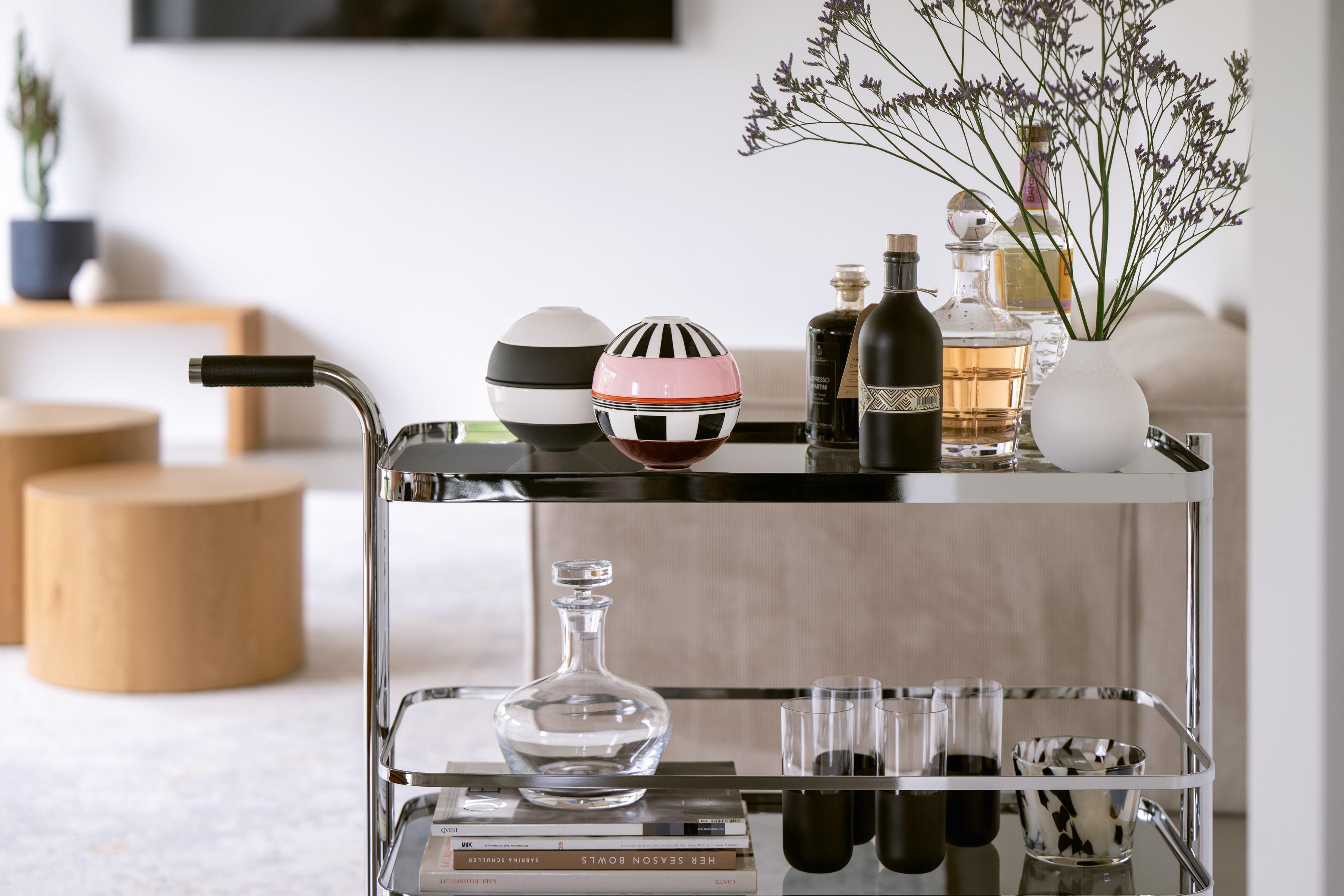 Servierwagen aus Chrom mit Karaffe, Gläsern, Spirituosen und kleinen Vasen. Im Hintergrund ein helles Wohnzimmer mit runden Holztischen und Sofalandschaft.