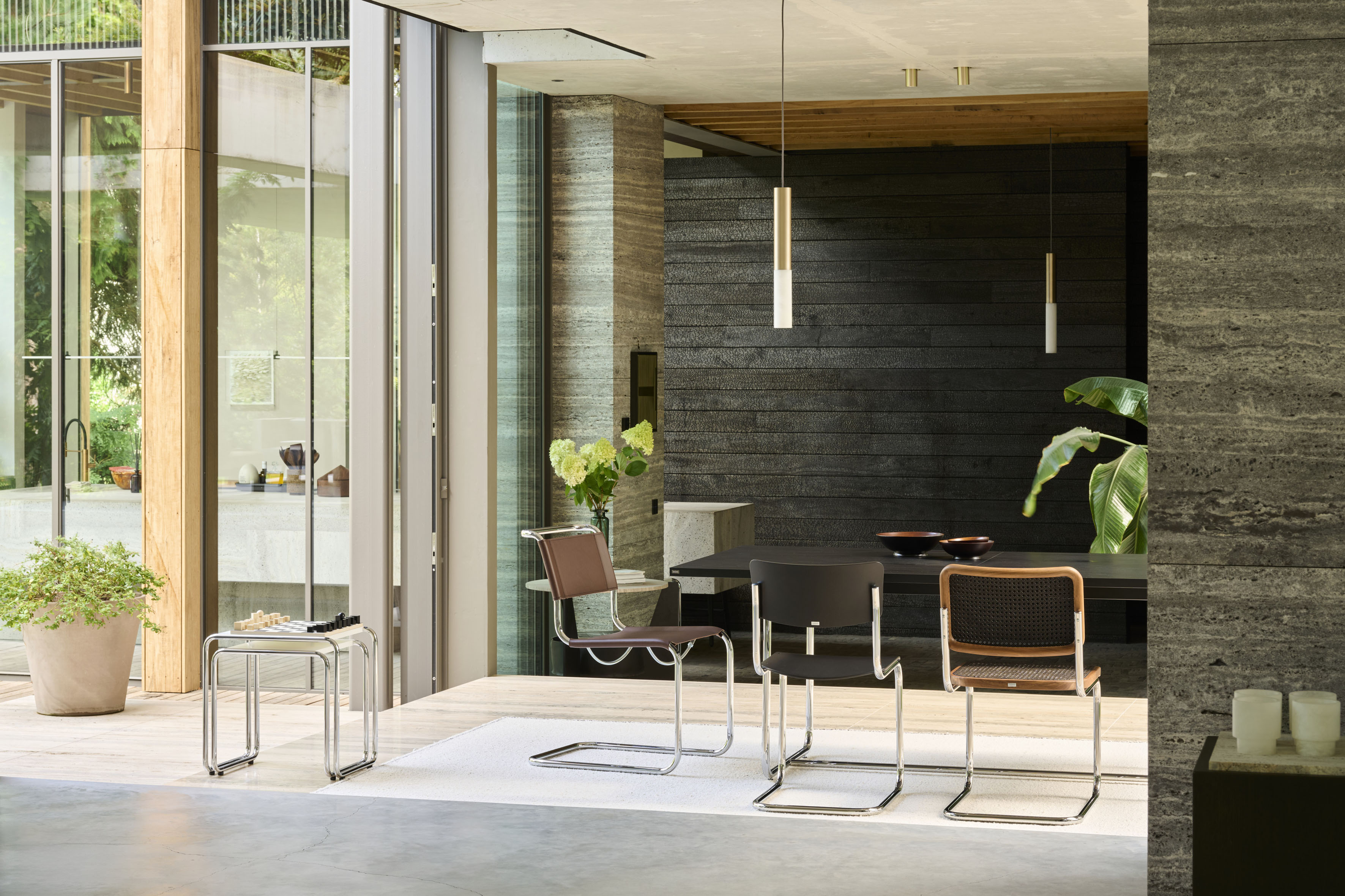 Moderner Essbereich mit drei Freischwinger Stühlen und zwei gläsernen Beistelltischen vor einer dunklen Holzstruktur Wand. Grosse Schiebetüren öffnen zum Garten, Pendelleuchten und viel Tageslicht schaffen eine ruhige, hochwertige Atmosphäre.
