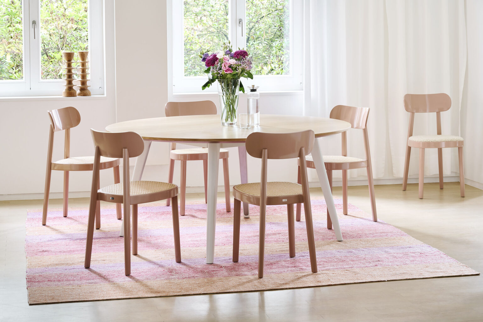 Heller Essbereich mit rundem Holztisch und sechs rosafarbenen Stühlen. Vase mit Blumen auf dem Tisch, rosa Teppich, weisse Vorhänge und Fenster im Hintergrund.