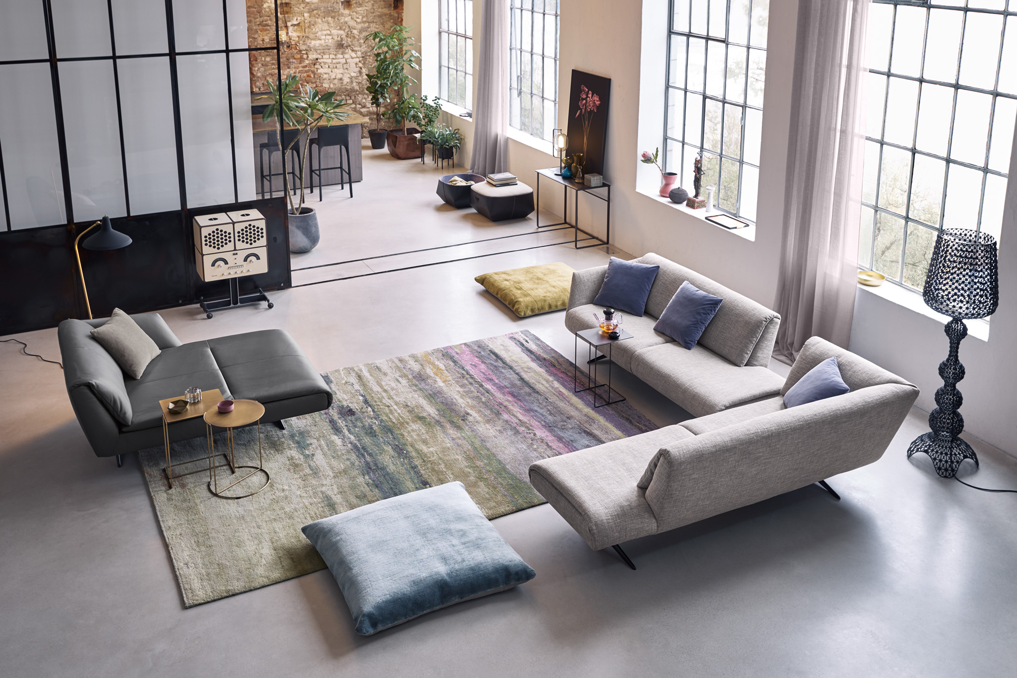 Wohnzimmer mit zwei modularen Sofas, farbigem Teppich und Bodenkissen. Grosse Sprossenfenster, Glasabtrennung, Beistelltische und mehrere Pflanzen sorgen für ein helles, wohnliches Ambiente.