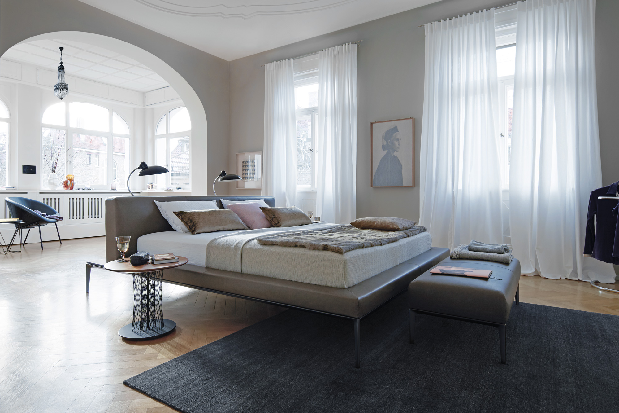 Helles Schlafzimmer mit hohen Fenstern und weissen Vorhängen, graues Polsterbett mit passender Bank am Fussende, Parkettboden und dunkler Teppich, runder Beistelltisch mit Drahtgestell, seitlich Arbeitsbereich in Nische mit Rundbögen, Wandbild mit Porträt und dezente Deko in neutralen Tönen.