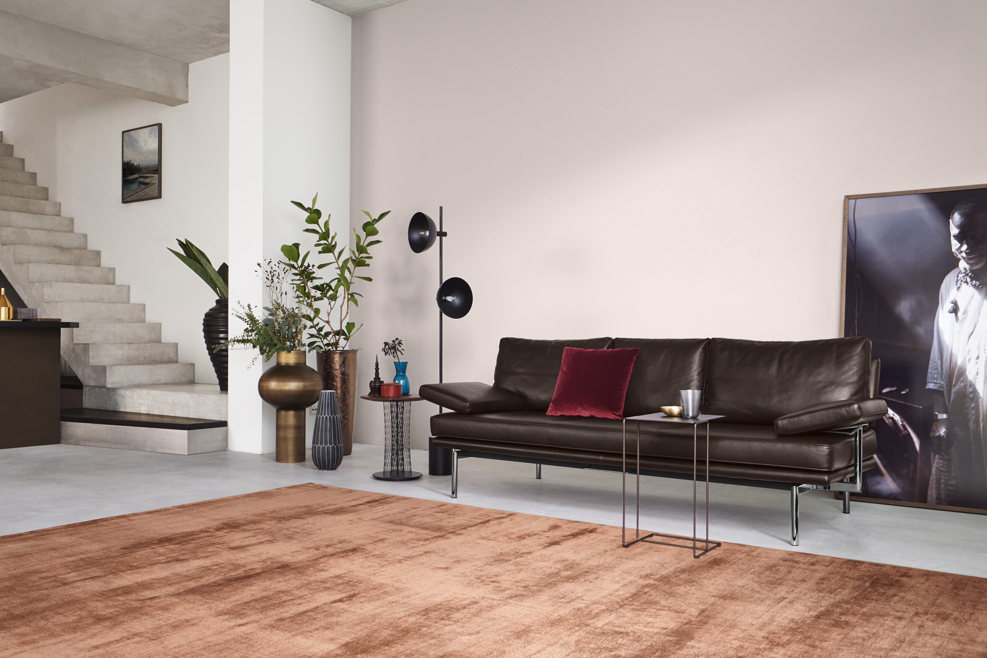 Minimalistisches Wohnzimmer mit dunkelbraunem Ledersofa auf filigranen Metallbeinen, rotes Samtkissen, zwei schlanke Beistelltische mit Becher, schwarze Stehleuchte mit runden Schirmen, grosse Pflanzen in skulpturalen Vasen, Betonwendeltreppe links, grossformatiges Porträt lehnend an der Wand, terrakottafarbener Teppich auf Sichtbetonboden.