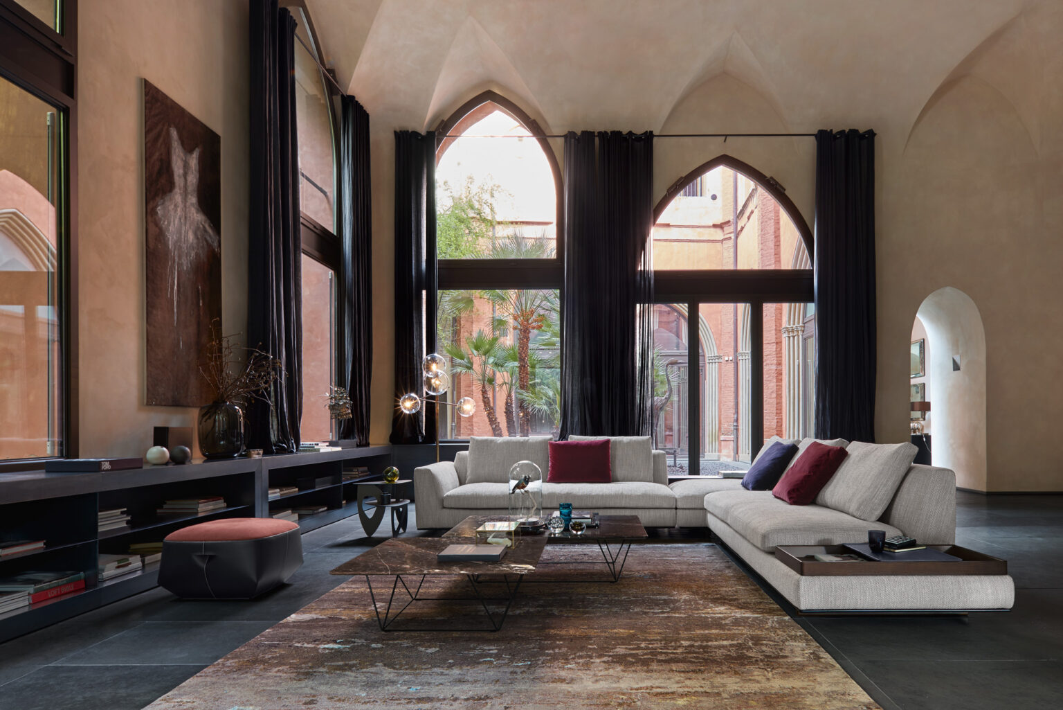 Grosszügiger Living Space mit hohen Spitzbögen und dunklen Vorhängen, helle modulare Sofalandschaft mit bunten Kissen vor raumhohen Fenstern zum Hof mit Palmen, niedrige Couchtische auf Vintage Teppich, lange Wandkonsole mit Deko und Kunstobjekten.