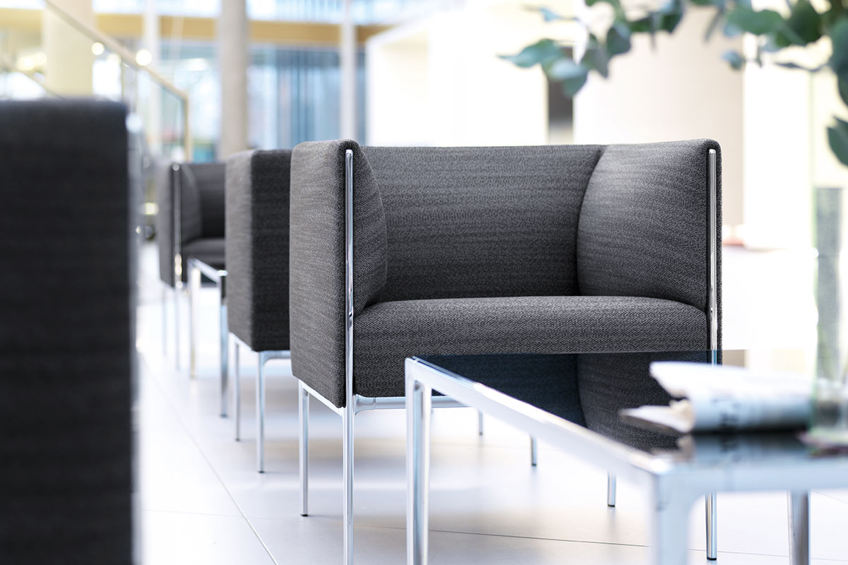 Moderner Wartebereich mit grauen kubischen Lounge Sesseln auf verchromtem Gestell, davor ein niedriger Glastisch mit Zeitung, helle Büroarchitektur im Hintergrund unscharf.