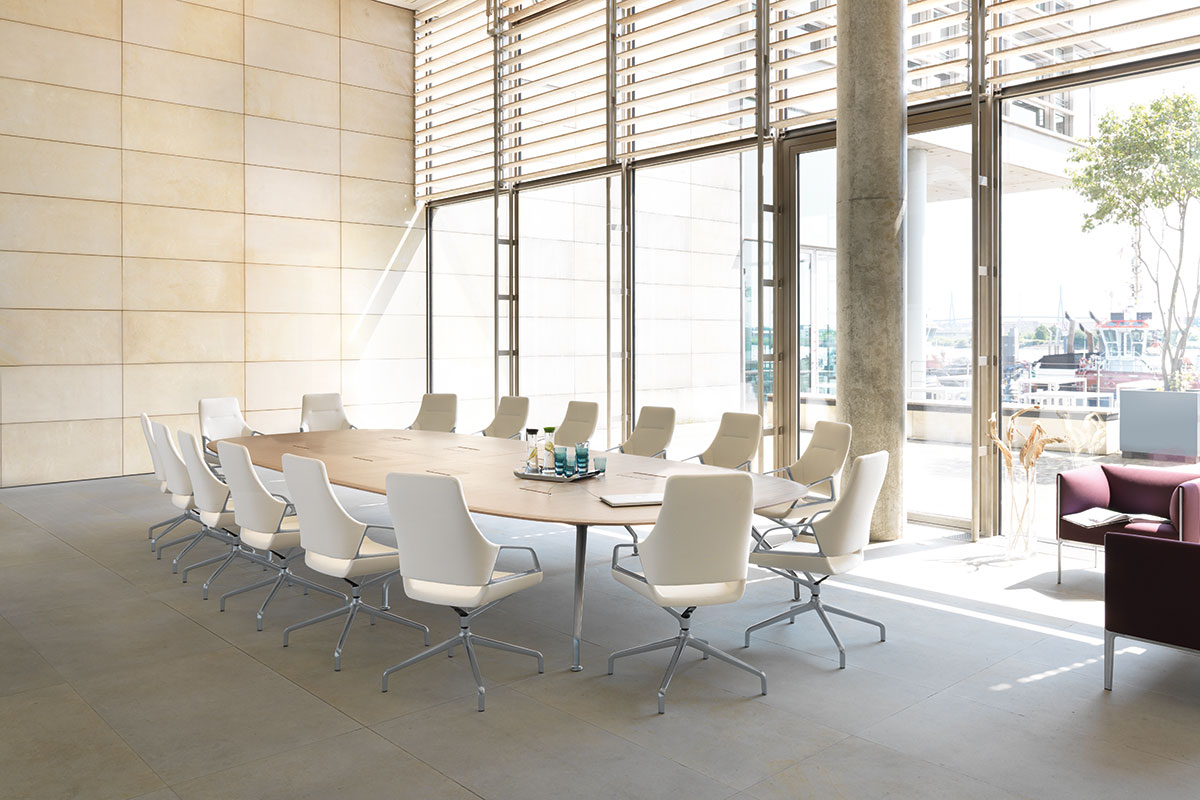 Heller Konferenzraum mit grossem ovalen Holztisch und weissen Drehstühlen, bodentiefe Fenster mit Lamellen bringen viel Tageslicht, rechts eine kleine Lounge und Ausblick auf den Aussenbereich mit Wasser und Booten.
