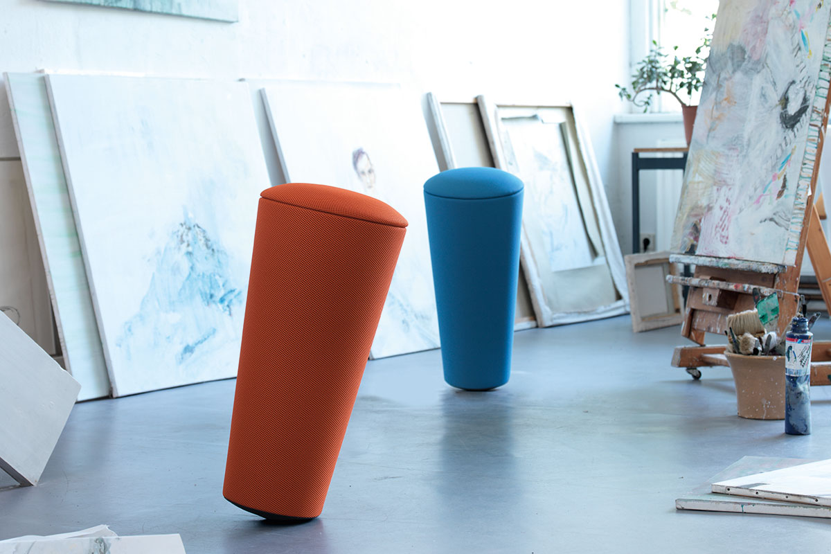 Zwei konische Balancehocker in Orange und Blau stehen in einem Künstleratelier zwischen angelehnten Leinwänden, Staffelei und Malutensilien auf grauem Boden.