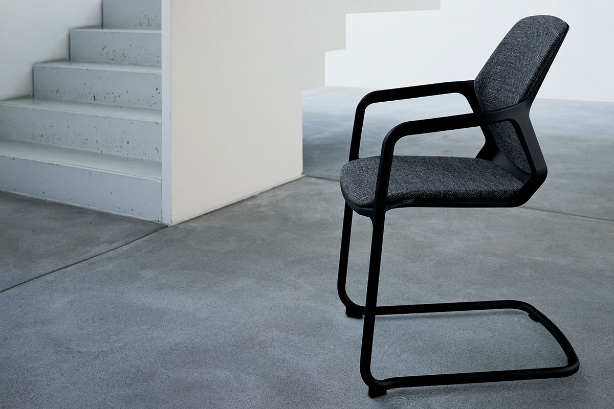 Freischwinger mit schwarzem Gestell und grauer Polsterung. Steht vor einer Betontreppe auf fugenlosem Betonboden. Minimalistische, monochrome Szene.