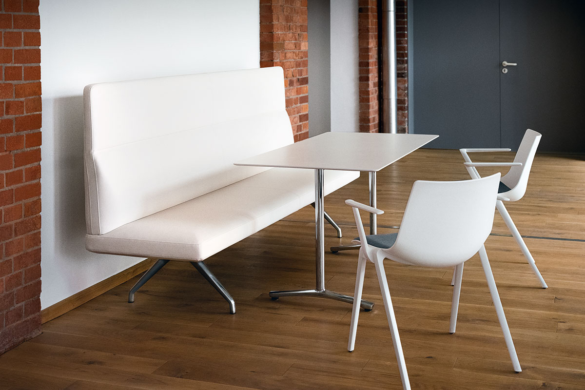 Heller Pausenbereich mit hoher, weisser Polsterbank auf Metallfuss, rechteckigem Tisch mit Chromgestell und zwei weissen Armstühlen mit grauen Sitzpolstern auf Parkett, Backsteinwand und graue Türe im Hintergrund.