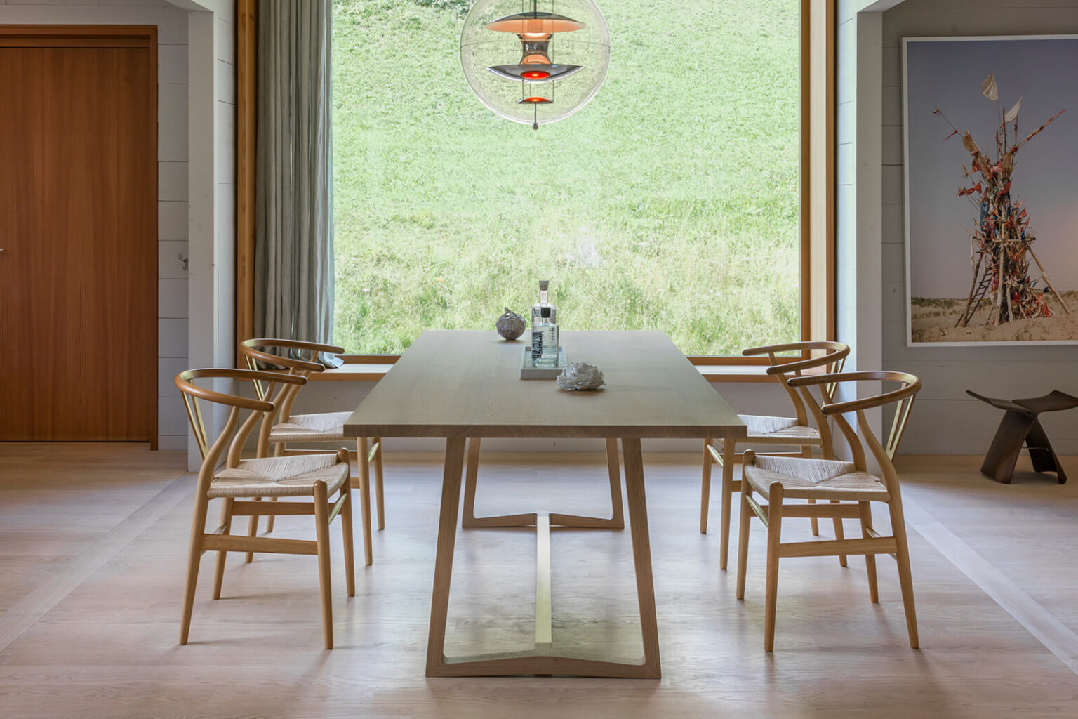 Helles Esszimmer mit grossem Holztisch und vier Wishbone-Stühlen vor bodentiefem Fenster mit Blick auf die grüne Wiese, darüber mehrteilige Pendelleuchte, rechts ein gerahmtes Wandbild.