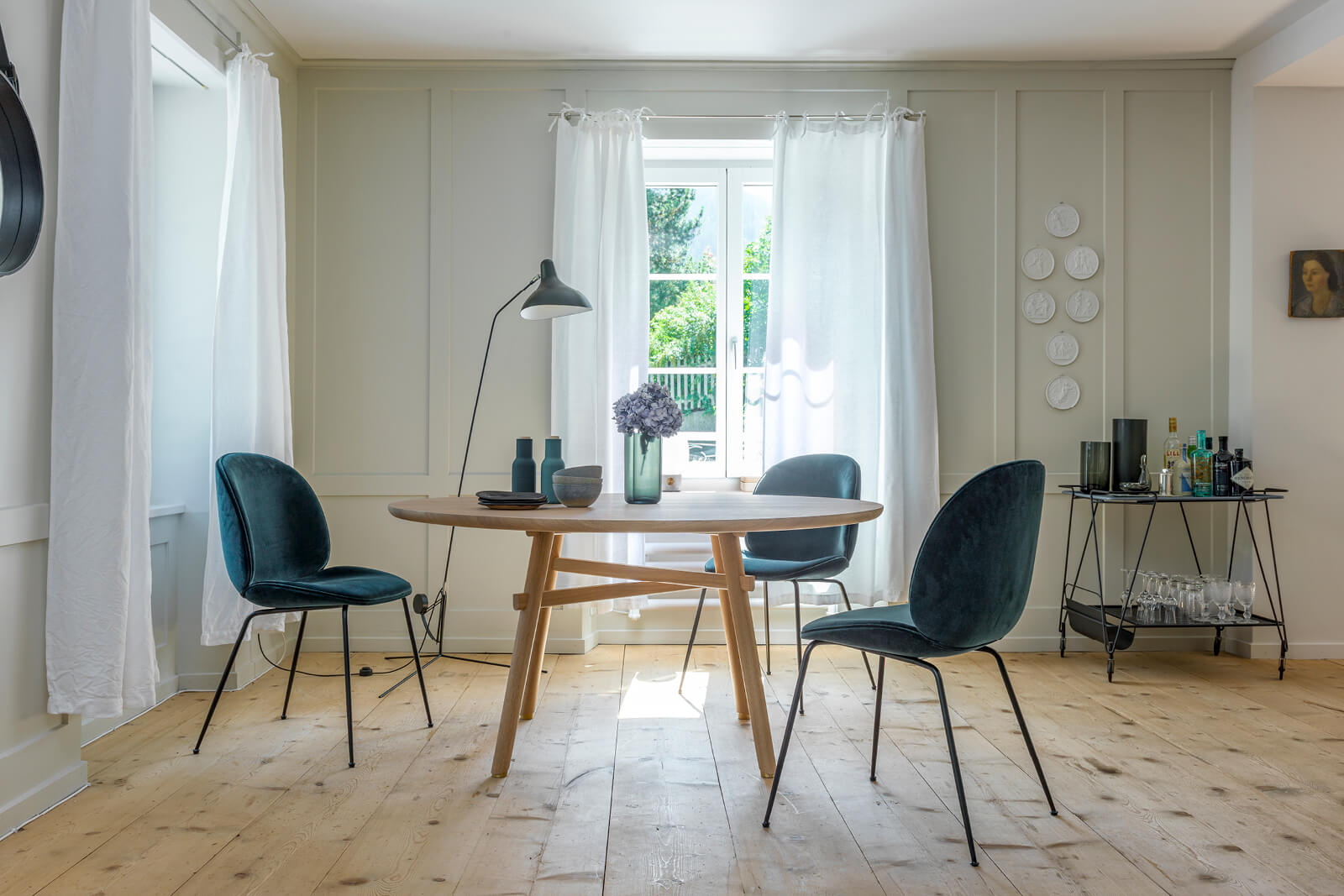 Heller Essbereich mit rundem Holztisch und drei samtigen, petrolfarbenen Stühlen, weisse Vorhänge vor einem Fenster, schwarze Stehleuchte links, Vasen mit Hortensie als Centerpiece, rechts Servierwagen mit Flaschen und Gläsern, Holzfussboden und Wandreliefs.