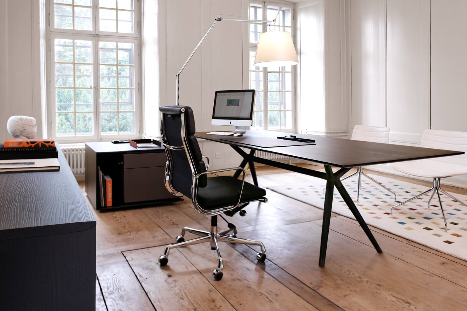 Helles Büro mit grossem dunklem Schreibtisch auf Stativbeinen, ergonomischem Drehstuhl mit Chromgestell, Rollcontainer und iMac auf Holzboden, hohe Sprossenfenster und Pendelleuchte, Besprechungsstühle im Hintergrund.