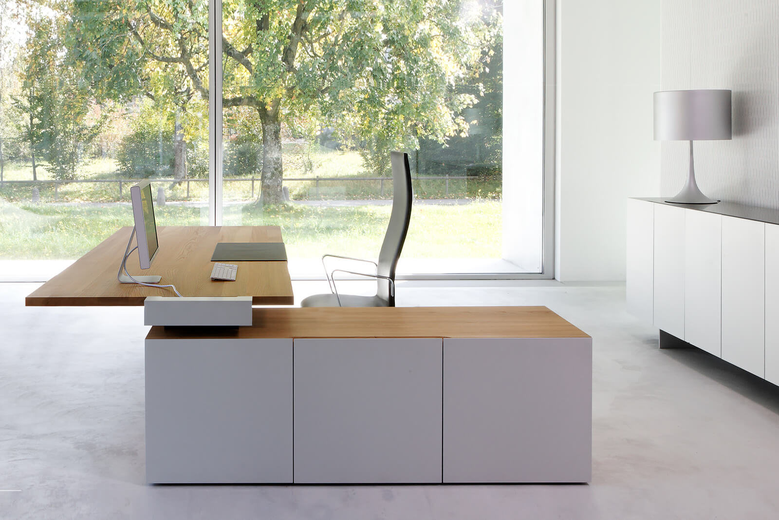 Lichtdurchflutetes Minimal-Office mit schwebendem Holztisch, Monitor und Tastatur, ergonomischem Drehstuhl und weissem Stauraummodul vor bodentiefem Fenster mit Blick ins Grün, rechts Sideboard mit Tischleuchte.