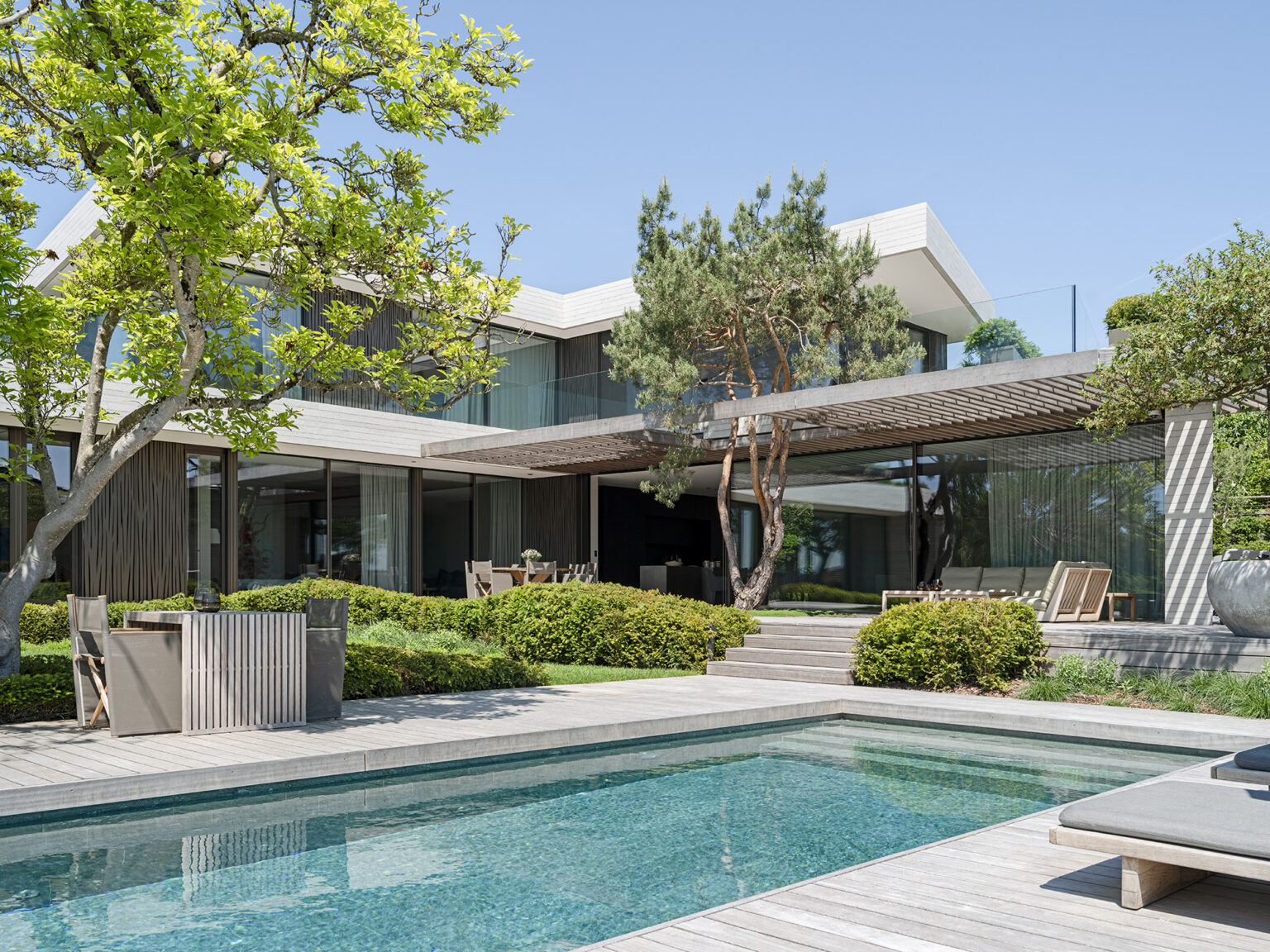 Modernes Privathaus mit Pool, Gartenanlage und weit auskragender Pergola vor grossen Glasfronten.