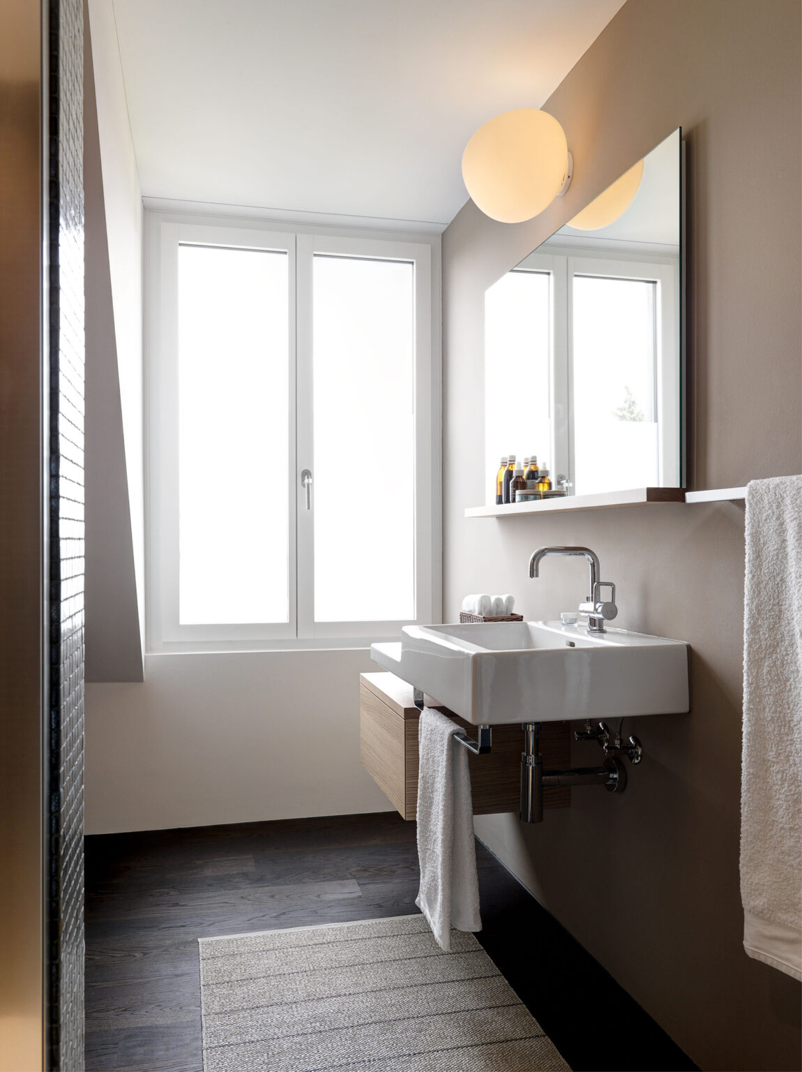 Helles Badezimmer mit Fenster, weissem Waschbecken, Spiegel und Kugel Wandleuchte.