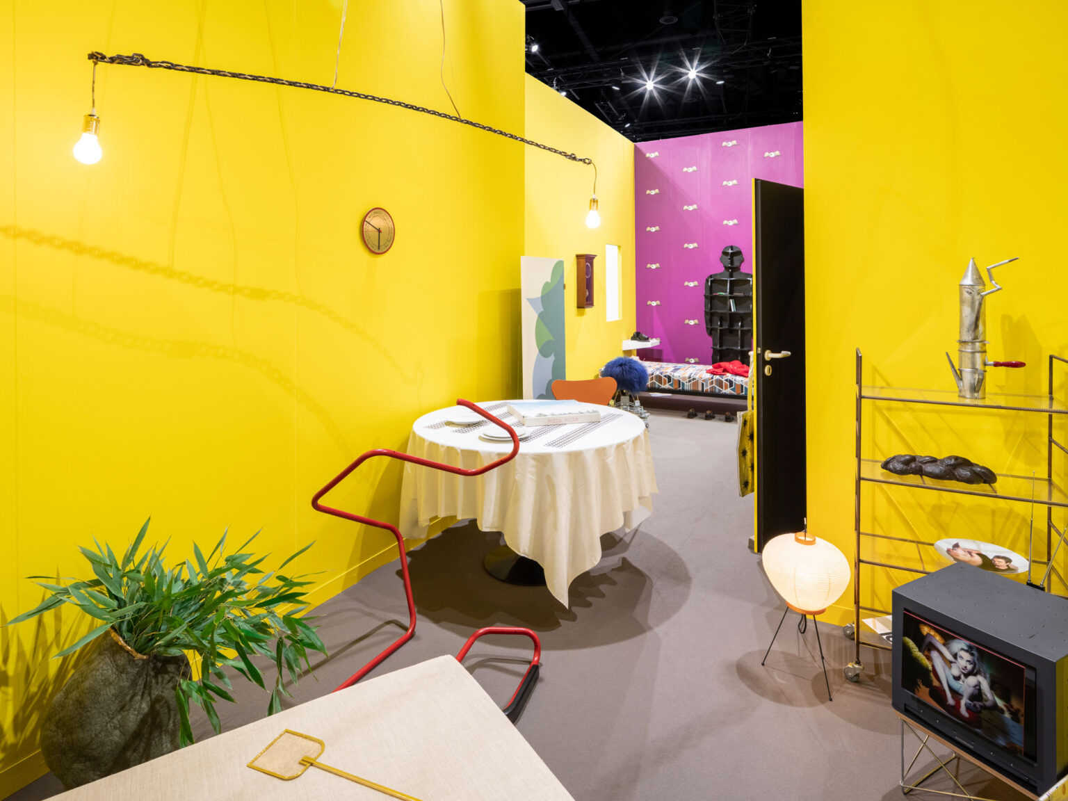 Gelber Ausstellungsraum mit rundem, weiss gedecktem Tisch, roter Rohrskulptur, Regal, Fernseher und offener Tür zu einem violetten Nebenraum.
