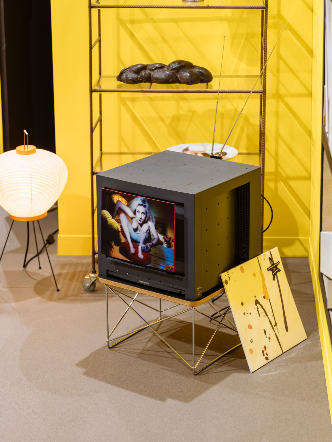 Schwarzer Röhrenfernseher auf filigranem Untergestell vor gelber Wand, daneben Regal, Kugelleuchte und dekorative Kunstobjekte.