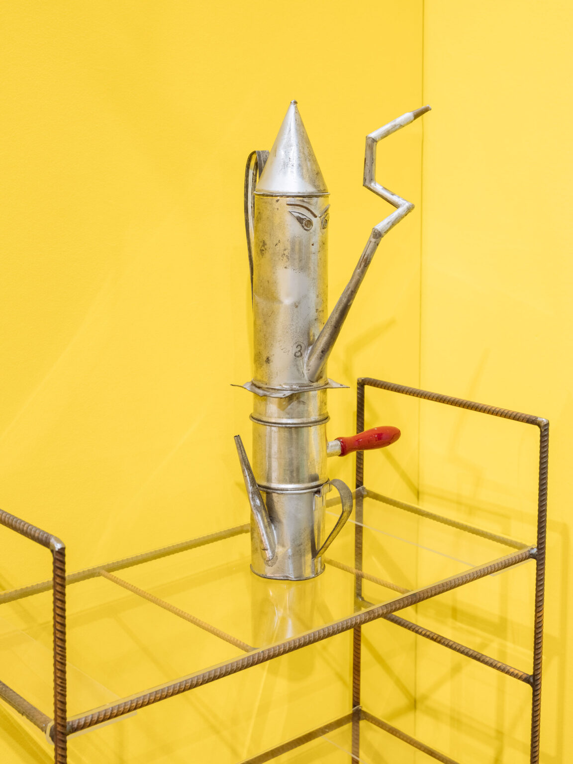 Silberfarbenes, skulpturales Metallobjekt mit rotem Griff auf einem offenen Regal vor leuchtend gelber Wand.