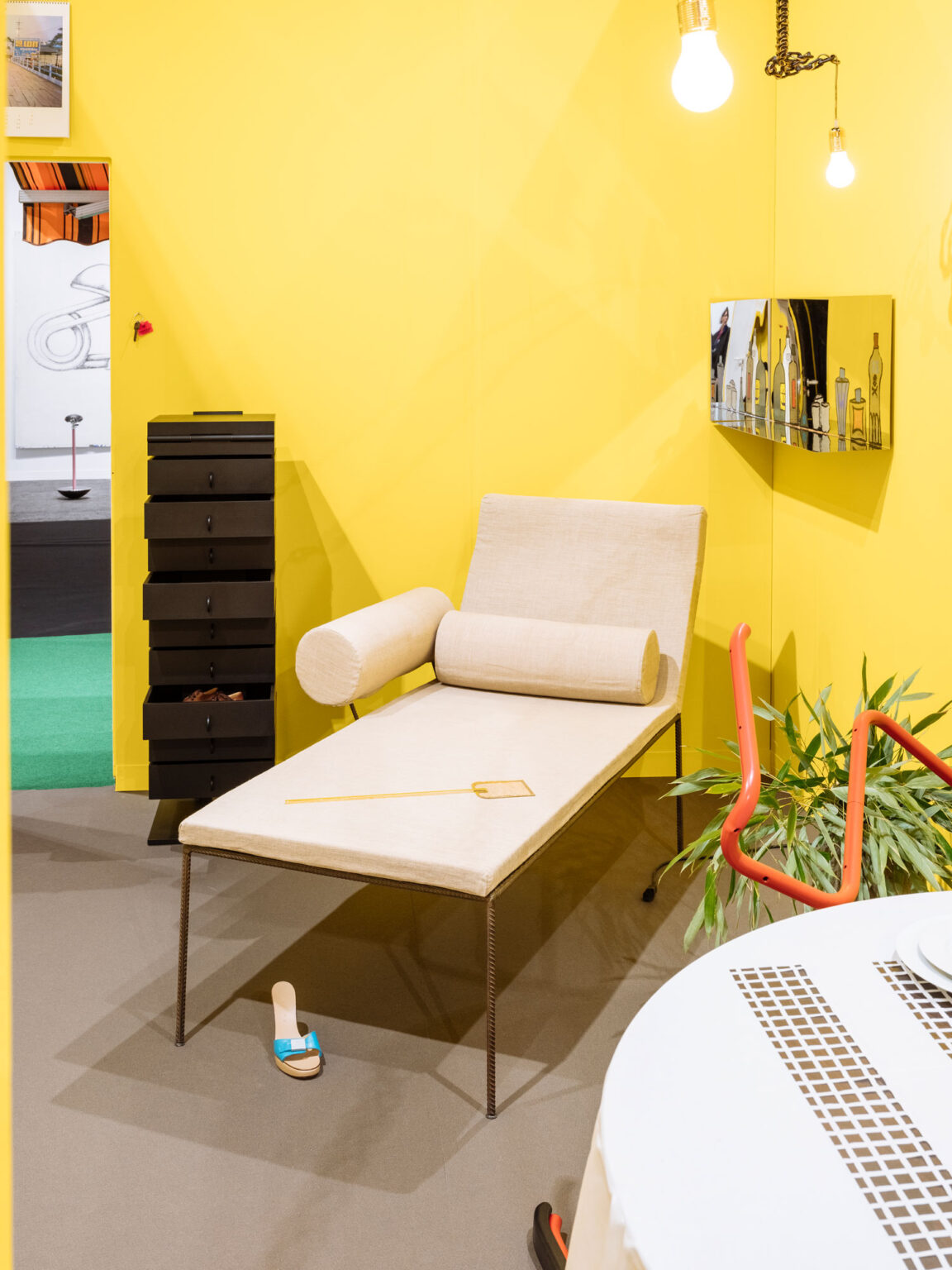 Beige Liege mit zwei zylindrischen Polstern vor gelber Wand, links ein schwarzer hoher Schubladenblock, rechts eine teilweise sichtbare rote Rohrskulptur, oben zwei Glühbirnen.