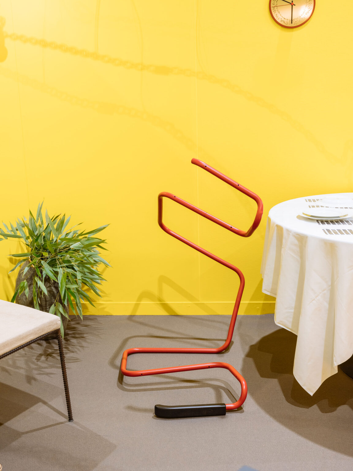 Rotes, s-förmiges Rohrobjekt vor gelber Wand neben einem runden Tisch mit weisser Tischdecke und einer Topfpflanze.
