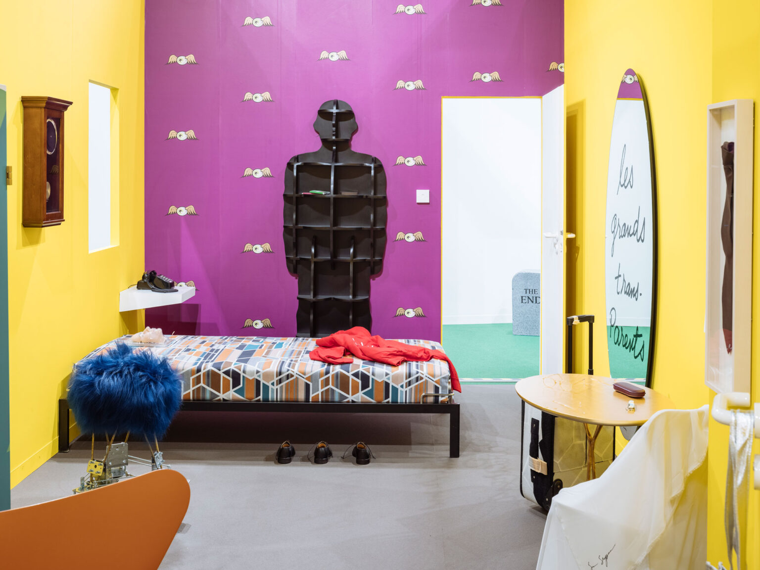 Gelber Raum mit violetter Akzentwand, Bett mit gemusterter Decke, schwarze menschenförmige Regalstruktur, offene Tür zu einem weissen Raum mit grünem Teppich.