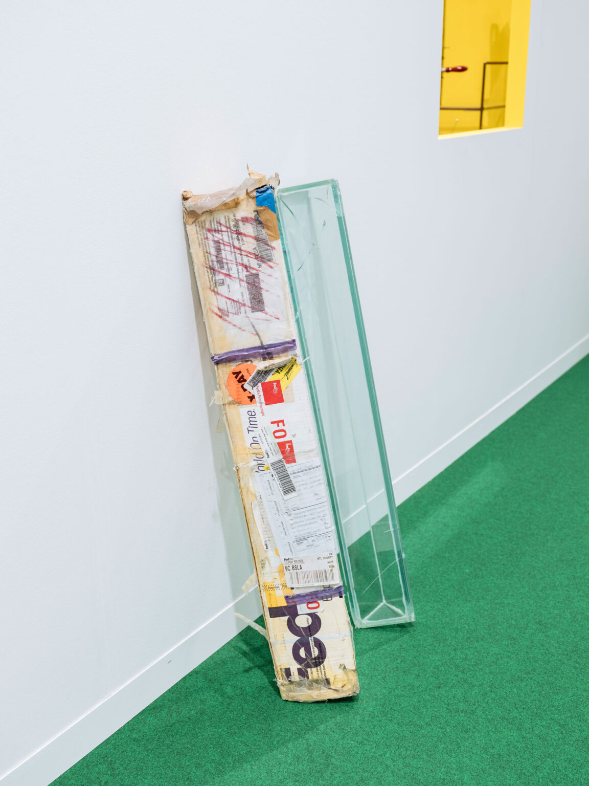 Grüner Boden vor weisser Wand, ein stark beklebtes FedEx Versandpaket lehnt schräg, daneben steht eine hohe, transparente Glasscheibe, oben rechts ein gelber Wandeinlass.