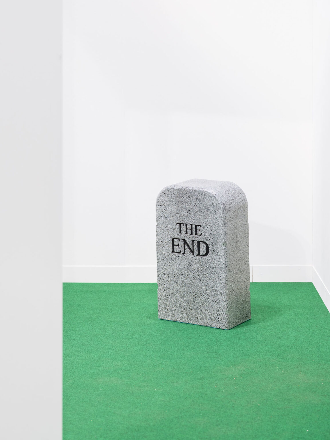 Steinblock mit der Aufschrift THE END auf grünem Boden vor weisser Wand.