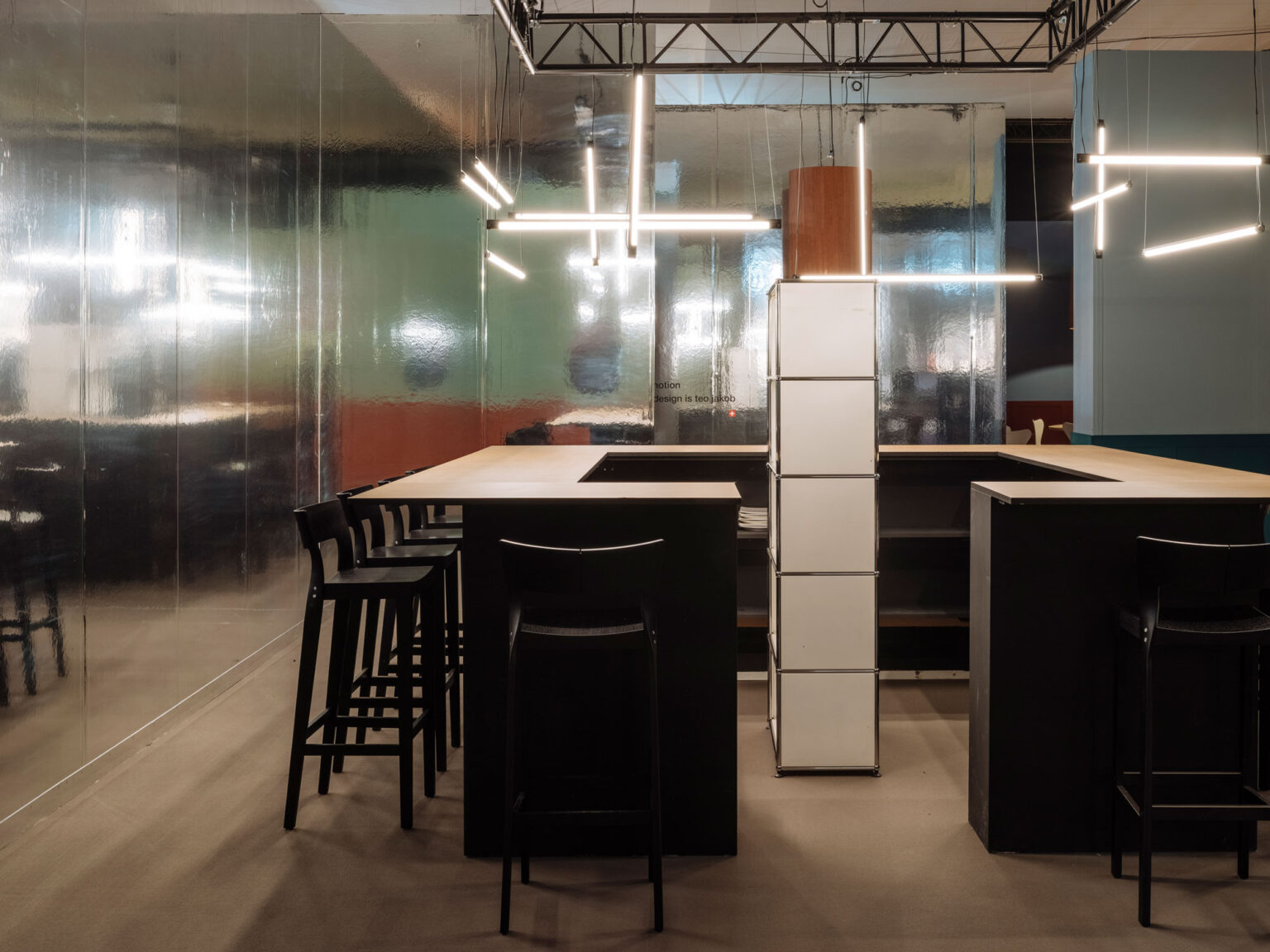 Offene Bar und Begegnungszone mit U förmigem Counter in Schwarz und Holz, schwarzen Barhockern, linearen Hängeleuchten und einer zentralen USM Säule. Spiegelnde Wandflächen verleihen der Artgeneve Konstellation Tiefe, Lichtreflexe und eine futuristische Raumwirkung.