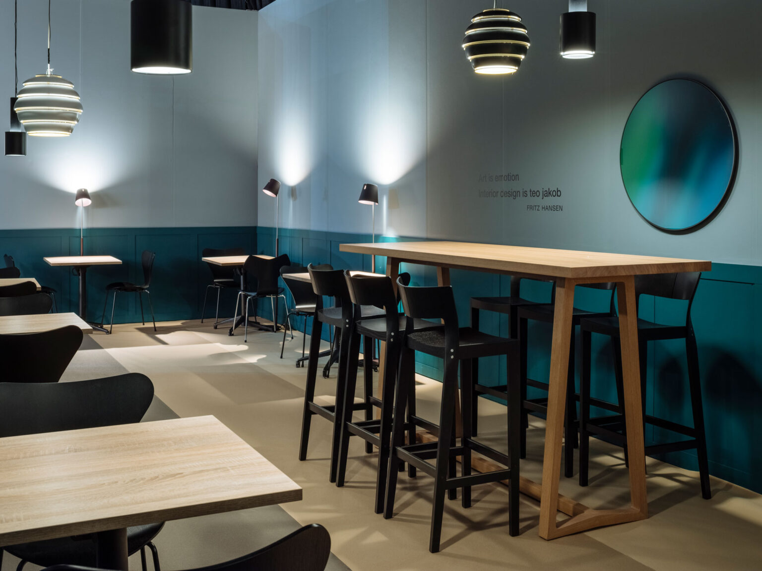 Lounge Setting mit hohem Holztisch und schwarzen Barhockern, kleinen Tischen, gedämpftem Licht und rundem Spiegel vor petrolfarbener Wand, Artgeneve Konstellation von teo jakob.
