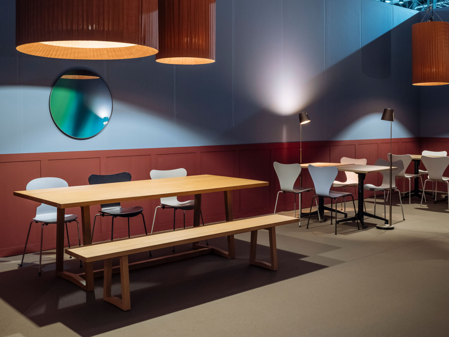 Lounge und Bistrobereich mit langen Holztischen, Sitzbank und verschiedenen Stühlen vor einer blau und bordeauxfarbenen Wand. Runde Spiegel und warme Pendelleuchten setzen prägnante Akzente.