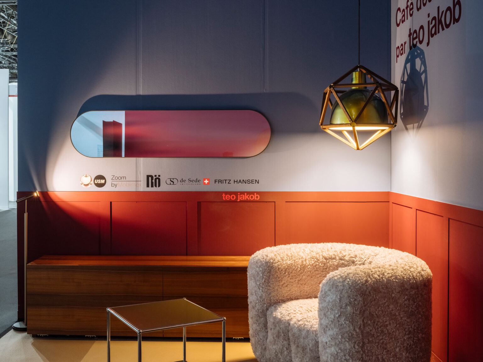 Brand Corner mit roter Wandvertäfelung, ovalem Farbspiegel und geometrischer Pendelleuchte. Partnerlogos und ein weicher Sessel runden die Szene ab.