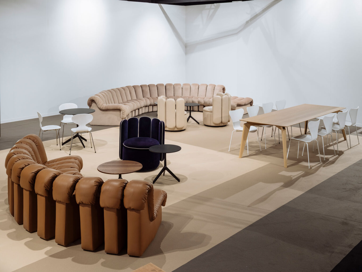 Lounge Bereich auf einer Kunstmesse mit modularen Sofas in Braun und Beige, runden Beistelltischen und einem langen Holztisch mit weissen Stühlen vor einer hellen Wand.