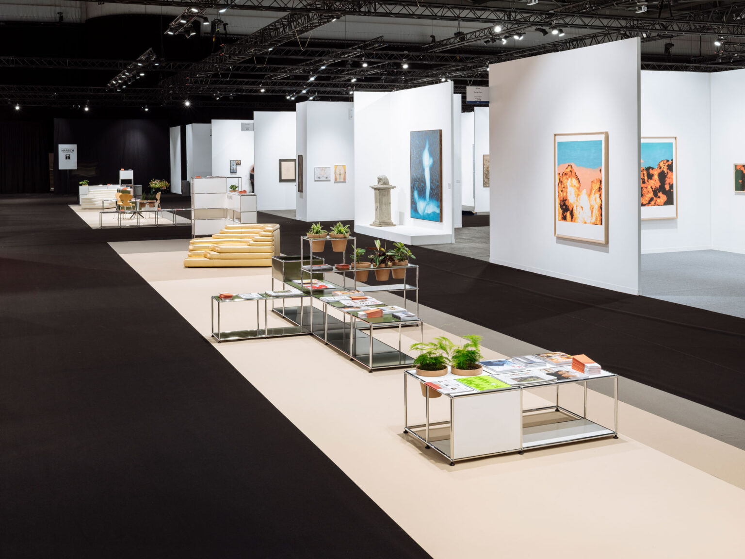 Blick durch einen Messestand mit USM Haller Modulen als niedrige Tische, darauf Magazine und Topfpflanzen, vor weissen Stellwänden mit gerahmter Kunst.