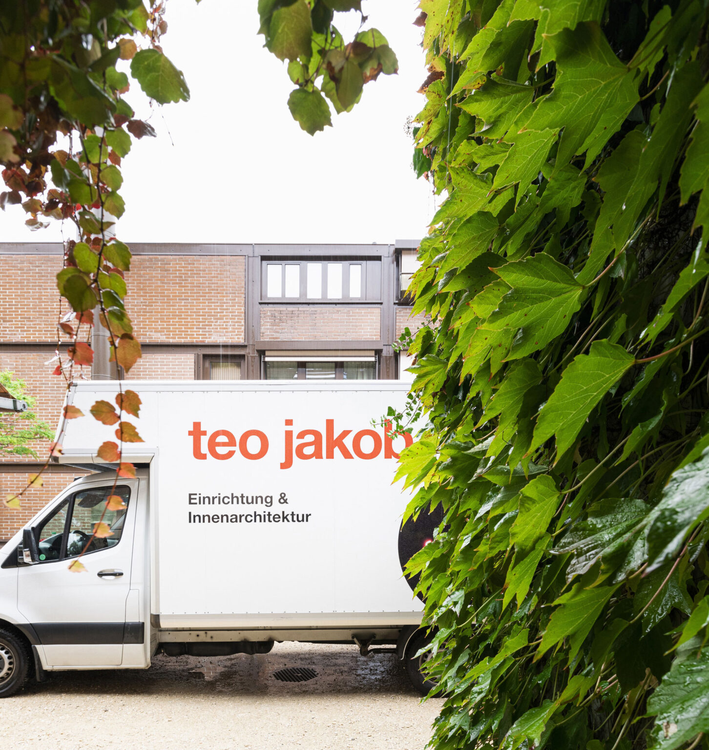Weisser Lieferwagen mit orangefarbigem teo jakob Logo und dem Schriftzug Einrichtung und Innenarchitektur, teilweise von grünem Efeu im Vordergrund verdeckt, vor einem Gebäude.