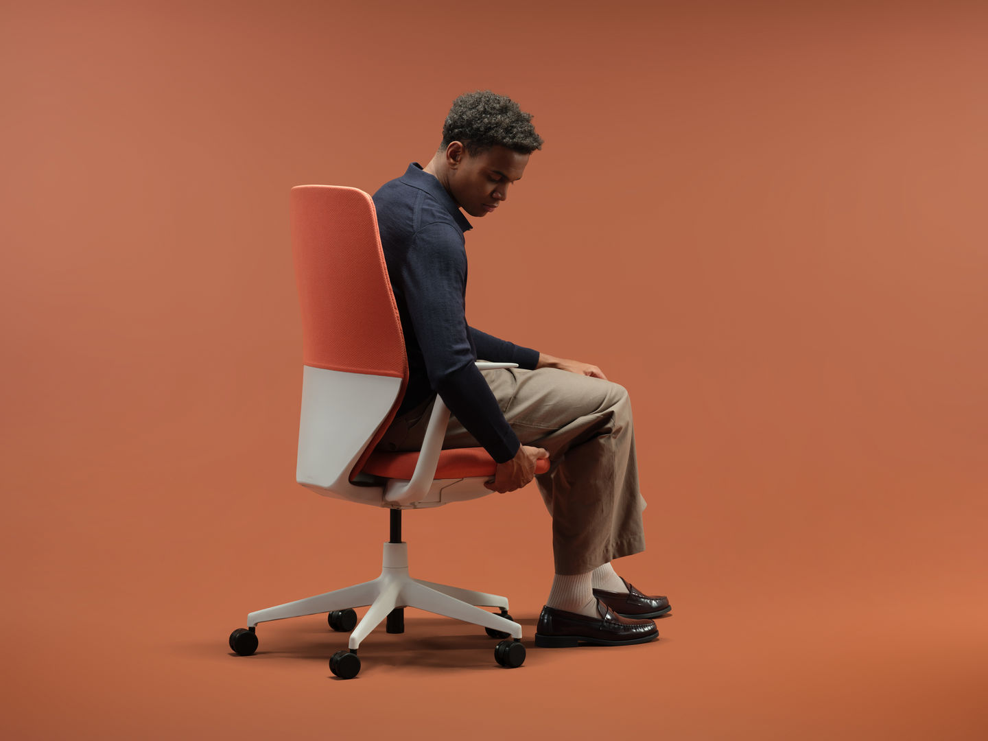 Person sitzt seitlich auf einem orangefarbenen Vitra Bürostuhl mit hoher Rückenlehne und weissem Drehgestell vor monochromem Hintergrund.